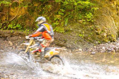 Sunny summer day. Enduro athlete overcomes a forest stream. Sunlight and lot of water sprayの写真素材
