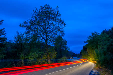 Tail lights illuminate an empty road in a night summer forest. Long winding trailsの写真素材