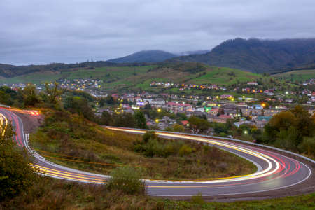 Cloudy evening on a mountain road. Headlight trails of fast moving cars. A small village shines with lights in the valleyの写真素材