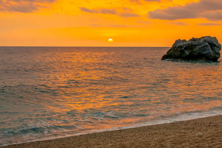 Lonely rock in the sea near the sandy beach. Colorful sunrise over the calm waters of the Greek Mediterranean coastの写真素材