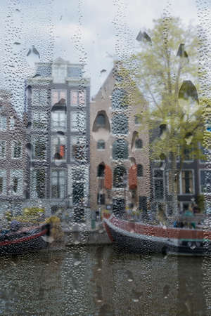 Netherlands. Canal embankment in Amsterdam in rainy weather from a high point. Residential barges and authentic Dutch houses. View through a window with raindropsの写真素材