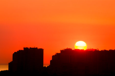 Dawn in the summer sky over the city. A large rising sun over the city roofs. Silhouettes of modern buildingsの写真素材