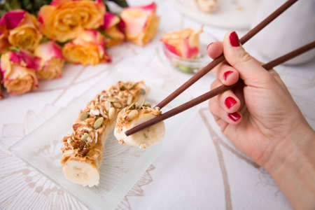 Banana sushi servered with nuts and granola on a glass plate. Hands with Japanese sticks. Woman eating healthy snack. Flowers and fruit breakfast or dessert.の写真素材