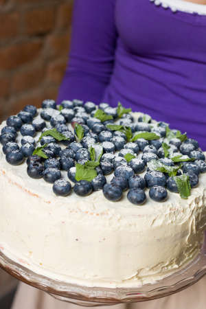 Woman in a lilac shirt holding a homemade white birthday cake with organic blueberries and mint on top. Party dessert. Celebration of birthday. Stylish cake with cream and berries.の写真素材
