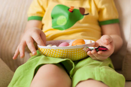 A small boy in a yellow t-shirt and green shorts sitting and eating Easter chocolate eggs. Easter...