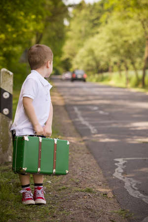 Adorable little kid boy in a white polo shirt and shorts holds a green retro suitcase and  stands on the countryside road waiting for a bus or a car. Ready for travel. Vacation time. Child on the roadの写真素材
