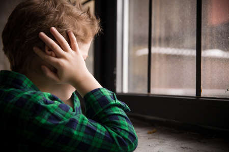 Little boy standing behind the window in sad mood. Sad Teenager looking in the Window and closing his ears with hands. Unhappy child in a plaid shirt. Alone at home. Upset.の写真素材