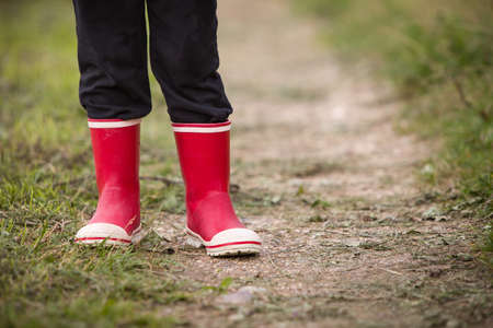 little kid boy in rain rubber boots. leg's shot. Child in bright red shoes. On a countryside road,...