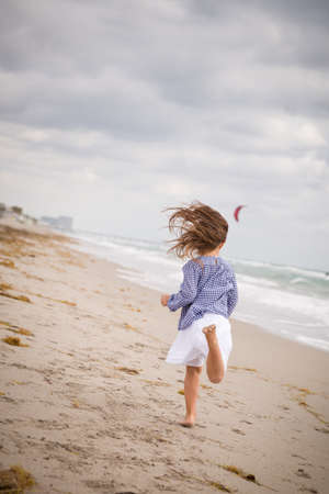 Little girl running on the ocean beach on a cloudy day. Vacation by the sea. Cute kid girl on the deserted beach. Summer, outdoors. Wind in the hair of a small girl. Beach landscape.の写真素材