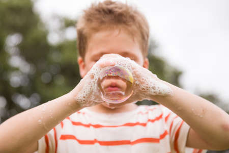 Portrait of adorable kid boy making soap bubble with his hands outdoors. Child blowing soap bubbles....