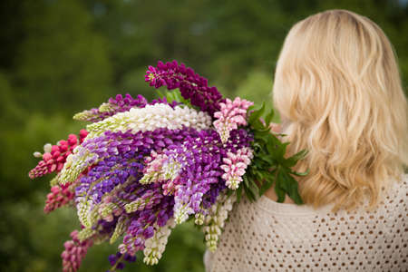 Back view on young girl holding a bunch of beautiful lupine flowers. Purple and pink lupin bouquet. Womanl with bouquet of wild  lupin flowers on sunny summer field.の写真素材