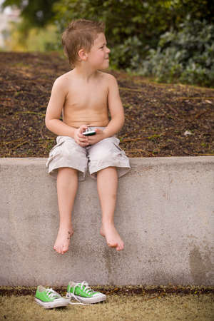 Cute little boy sitting in the park and taking photo with smartphone in a sunny summer day. Outdoors. Child with telephone playing games  and having fun on the sun. Children and technology.の写真素材