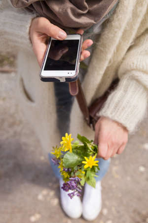 woman holding a beautiful bunch of wild spring flowers and taking photo of it with her smartphone. Bouquet of yellow and blue flowers in hands. Making picture by the telephone.の写真素材