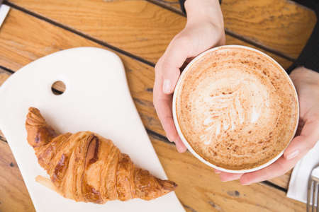 Top view on woman's hand holding big cup with cappuccino coffee and fresh French croissant on a...