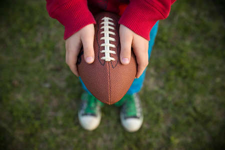 Top view on hands of little kid boy holding football on the stadium on a sunny day. Child  ready to throw a football. Sport concept. Sport activities for children outdoors.の写真素材