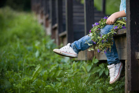 Closeup of boy in white sneakers sitting on wooden stairs and holding a nice summer flower wreath in his hands. Blue summer flowers in child's hands. Selective focus.の写真素材