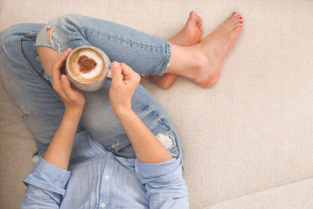 Woman sitting on the sofa and drinking coffee at home. Closeup of woman hands holding cup of coffee...