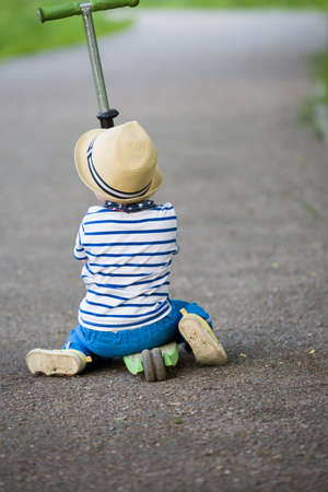 Back view on a cute toddler boy in straw hat sitting on a scooter on the road in the park. Child...