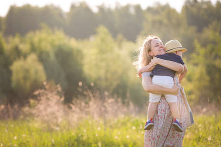 Happy family walking in the park on a summer sunny day. Mother hugging her toddler boy on a meadow on sunset. Mum with son. Woman with cute child outdoors.の写真素材