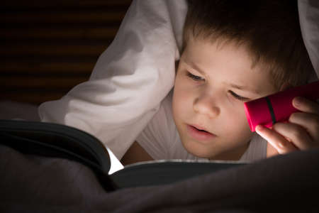 Small kid boy with a smile hiding under a blanket and reading a book with his flashlight in his bedroom at night. Enjoying learning and books.の写真素材