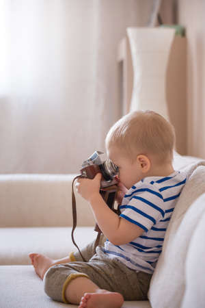 Cute adorable toddler boy sitting on the sofa in the living room and playing with vintage photo camera. Child taking picture with old camera. Future photographer. Kid with retro camera.の写真素材