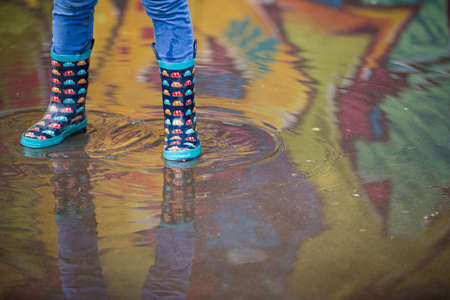 Kid boy in funny rubber boots standing in the puddle in the street after rain. Pair of  colorful rubber boots in a big puddle with graffiti reflections. Boy having fun after rain. Outdoor.の写真素材
