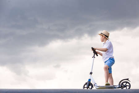 Cute kid boy riding his scooter into cloudy sky - happiness, childhood, freedom, speed and active lifestyle concept - smiling boy in straw hat on scooter in air over grey rainy sky.の写真素材