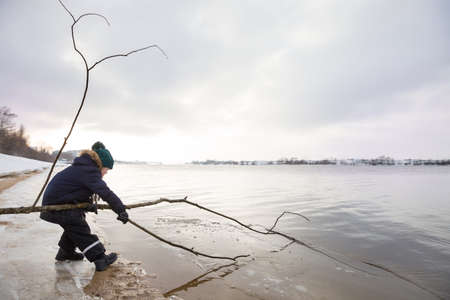 Little boy in winter cloths playing with tree stick and ice on the beach. Nature park and outdoors. Playing child at the river shore. Young explorer by the water. spring in the city. Ice in the water.の写真素材