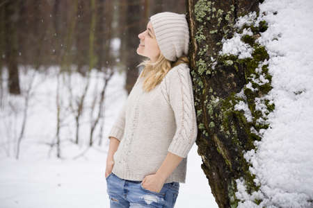 Young blond woman in a warm sweater and a hat standing near a tree in the forest. Winter walks outdoors. Snow in the forest.の写真素材