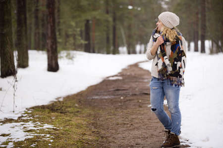 Young blond girl in blue jeans, warm sweater and bright scarf standing in a winter forest. Winter walks outdoors.の写真素材