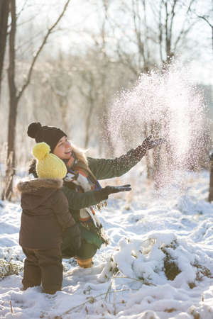 Happy family having fun with snow in the park on a sunny day. Mother with cute toddler child enjoying winter outdoors. Mom with boy. Winter and holidays seasons concept.の写真素材