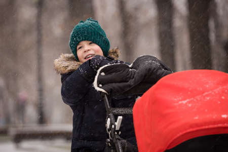 Portrait of cute happy kid boy in winter clothes pushing stroller with his siblling. Child with baby carriage outdoors on a cold day. Family conceptの写真素材