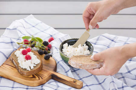 Woman taking organic farming cottage cheese from green bowl. Breakfast: slice of whole wheat bread with Homemade Ricotta cheese served with raspberries and blackberries on wooden board on linen fabricの写真素材