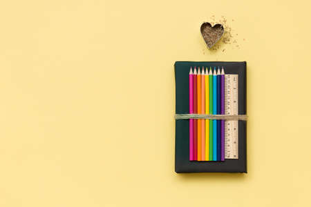 Colorful school and office supplies  - pencils and ruler on black book with shavings in shape of heart on light yellow background. Top view with copy space.の写真素材