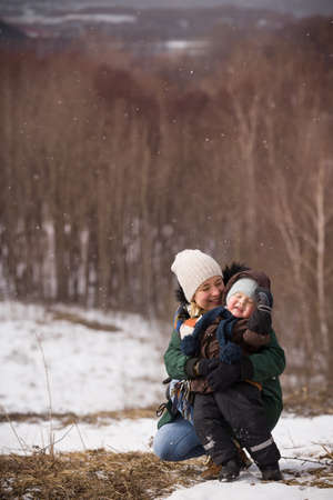 Beautiful woman with her small adorable toddler boy sitting on the ground in a winter day.  Forest background. Family walking in the city park. Happy kid and his mom.の写真素材