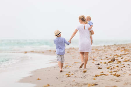 Young mother in white dress walking with her two little boy along the ocean beach. Woman with a baby and a boy enjoying vacation by the sea. On the empty beach. Motherhood.の写真素材