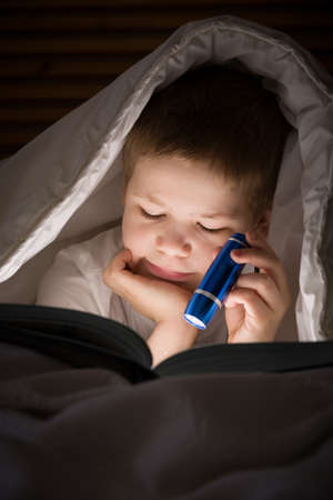 Small kid boy with a smile hiding under a blanket and reading a book with his flashlight in his bedroom at night. Enjoying learning and books.の写真素材