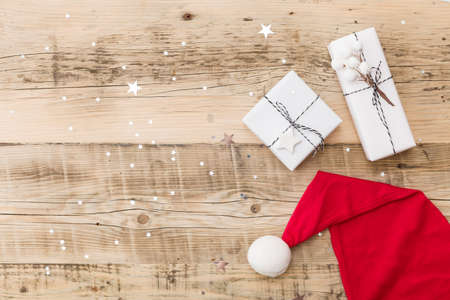 Top view on nice Christmas gifts wrapped in white gift paper, Christmas tree decorations in Santa's hat on wooden background with sparkling stars. New Year, holidays and celebration conceptの写真素材