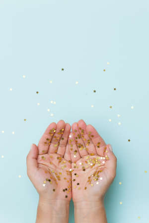 Top view on woman's hands covered with golden stars confetti on light blue background. Christmas and...