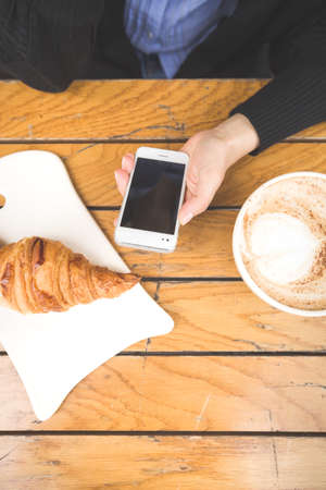 Top view on woman's hands holding big cup with cappuccino coffee and fresh French croissant on a wooden table in outdoor cafe. Breakfast with coffee and bread.の写真素材