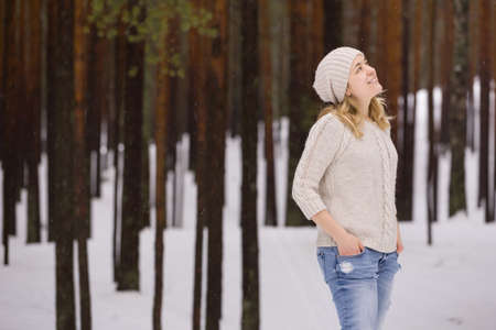 Young blond woman in a warm sweater and a hat standing in the forest, smiling and looking into sky. Winter walks outdoors. Snow in the forest.の写真素材
