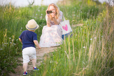 Mother taking photo of her adorable little toddler boy with smartphone. Family walking in the park. Small child with mom.の写真素材