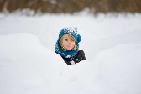 Portrait of adorable little kid boy with long blond hair playing with snow outdoors. Child with blue scarf and hat walking and having fun on a windy winter day.の写真素材