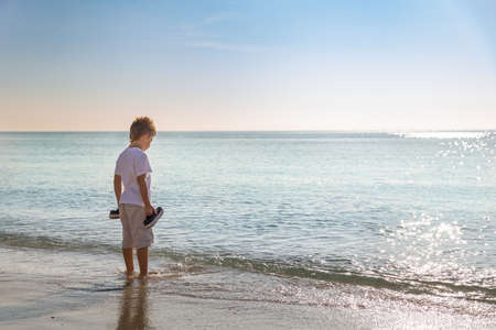 Adorable kid boy standing on the sandy beach by the ocean and looking on the gorgeous summer view. Miami summer vacations. Blue sky with clouds as a background. Child on amazing beach.の写真素材