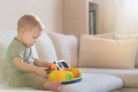 Cute blond toddler boy sitting on the sofa in the living room and playing with a toy. Early learning for kids. Activities with children at home.の写真素材