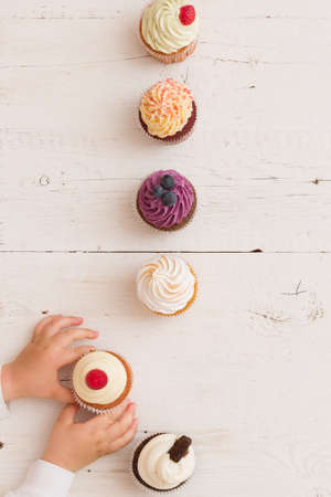 Top view on homemade vanilla, blueberyy, lemon, chocolate cupcakes in row and child's hands on white retro wooden background. Boy eating cupcake.Healthy food, snack for birthday party or celebrationの写真素材