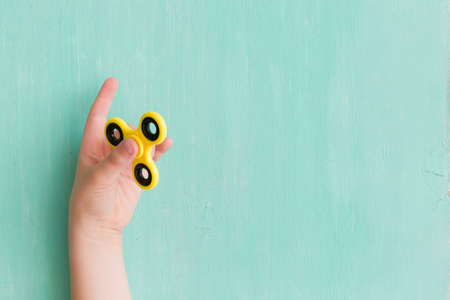 Child's hand spinning a fidget spinner device on turquoise background. Top view. Playing with a yellow hand spinner fidget toyの写真素材