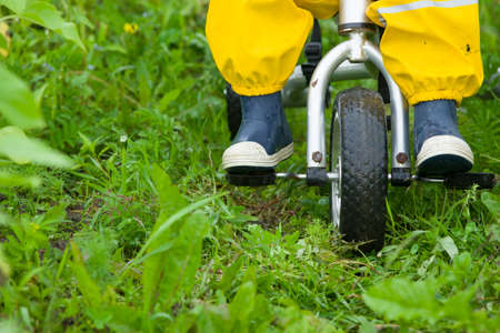 Closeup of toddler child feet in rubber boots and pants riding a bicycle on green grass. Boy walking outdoors after the rain.の写真素材