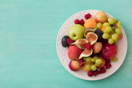 Top view on assortment of juicy fruits on white plate and turquoise wooden table background. Organic raspberries, apricots, apples, figs,plums, grapes - summer dessert or snack. healthy eating conceptの写真素材