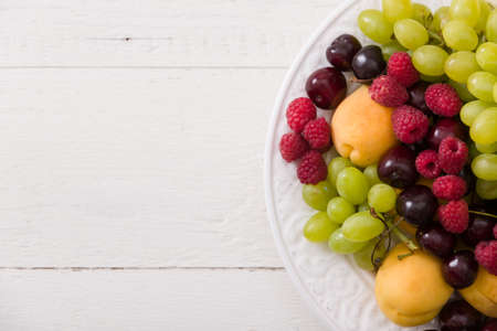 Top view on assortment of juicy fruits on white plate and white table background. Organic raspberries, apricots, melon, cherries - summer dessert or snack. healthy eating concept.の写真素材